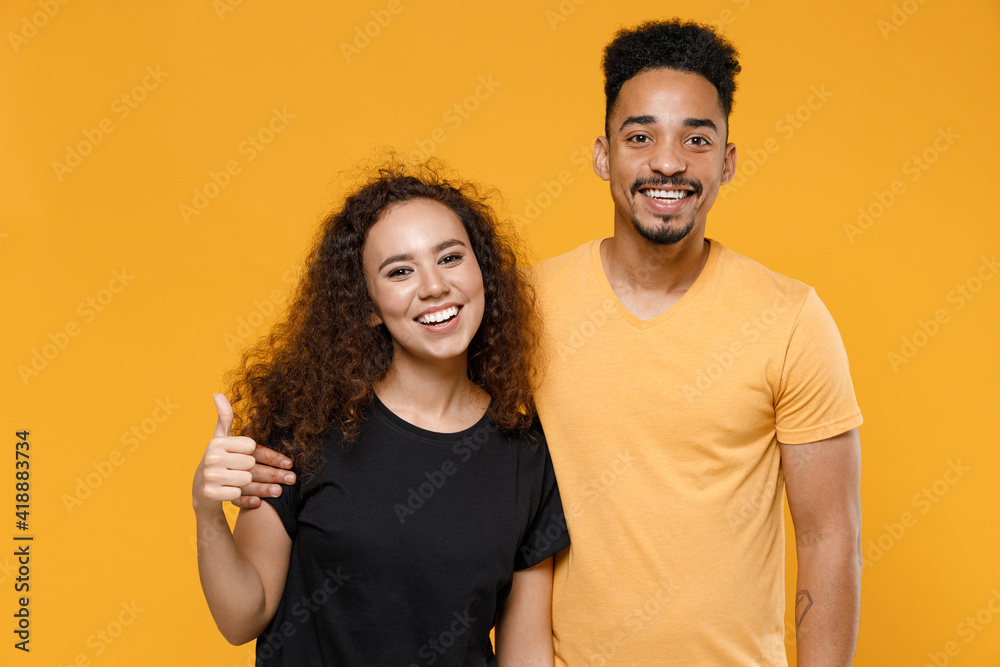 Fototapeta premium Young couple two american friends family happy satisfied smiling african man woman 20s together in black tshirt hug girlfriend show thumb up like gesture isolated on yellow background studio portrait