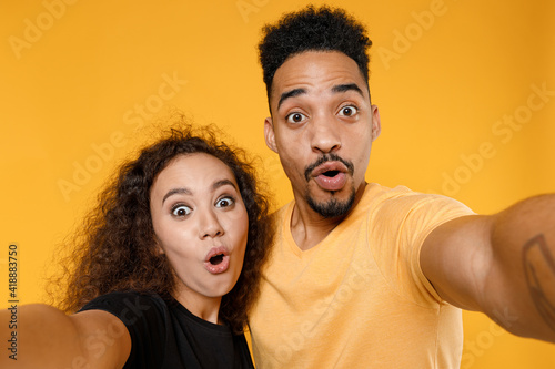 Close up young couple two friends together family surprised shocked impress african man woman in black t-shirt do selfie shot on mobile phone look camera isolated on yellow background studio portrait.