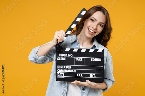 Young smiling happy brunette positive fun woman 20s in stylish denim shirt white t-shirt holding classic black film making clapperboard look camera isolated on yellow color background studio portrait.