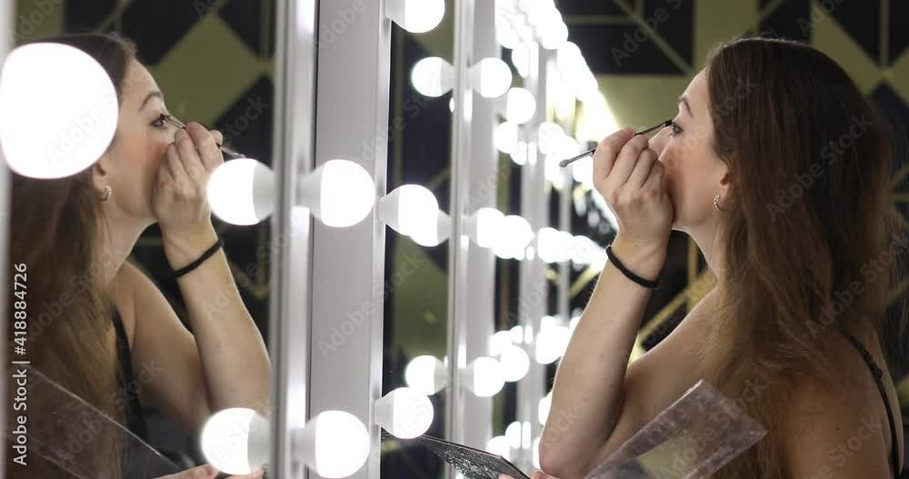 Beautiful young woman doing makeup in the dressing room in front of the mirror. Model uses a brush to apply shadows on the eyelids of the eyes