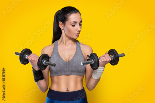 Young sportive attractive caucasian athlete doing light weight lifting in grey top and blue leggings - Studio shooting with pretty brunette posing during gymnastics workout weights routine