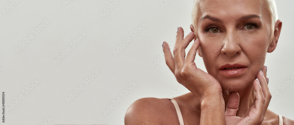 A skin care routine after 50. Attractive caucasian mature woman in underwear touching her face and looking at camera while posing against light background - obrazy, fototapety, plakaty 