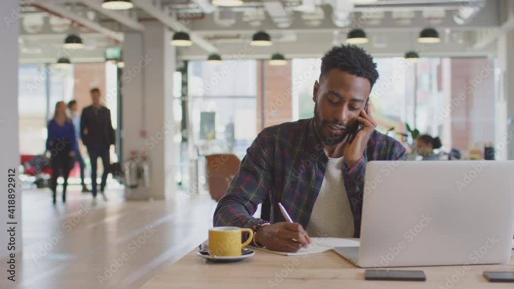 Dolly shot of casually dressed young businessman laughing on phone call sitting at desk in modern open plan start up office with colleagues in background - shot in slow motion