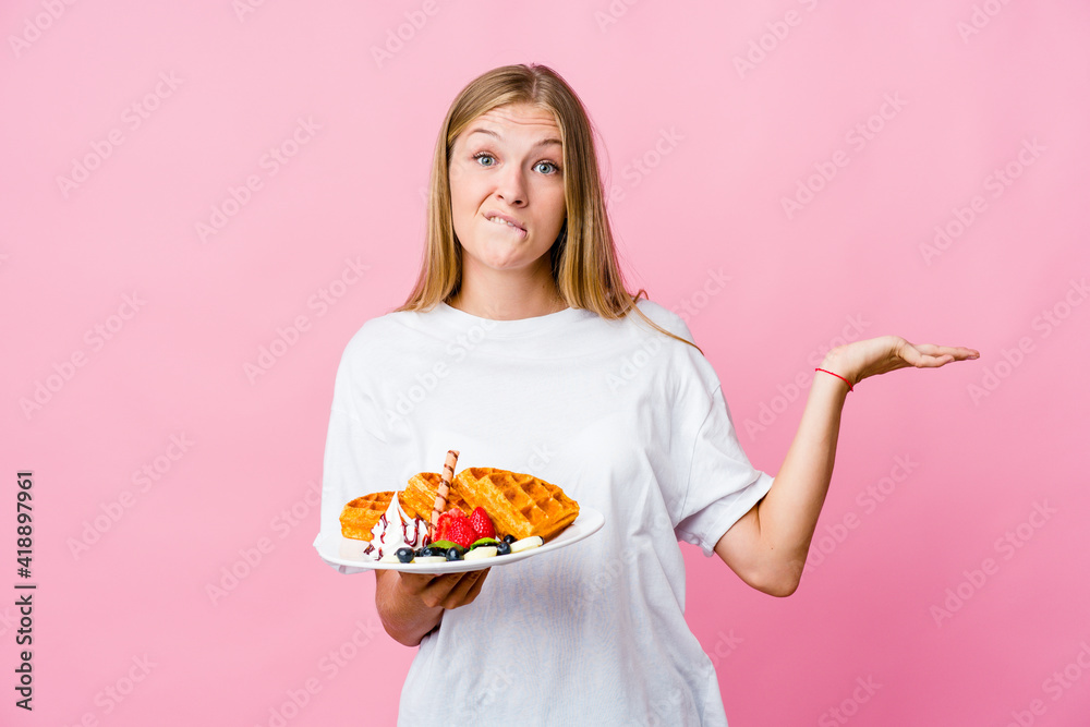 Young russian woman eating a waffle isolated confused and doubtful shrugging shoulders to hold a copy space. Young russian woman eating a waffle isolated confused and doubtful shrugging shoulders to hold a copy space.