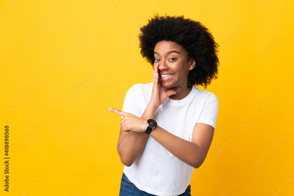 Young African American woman isolated on yellow background pointing to the side to present a product and whispering something Young African American woman isolated on yellow background pointing to the side to present a product and whispering something
