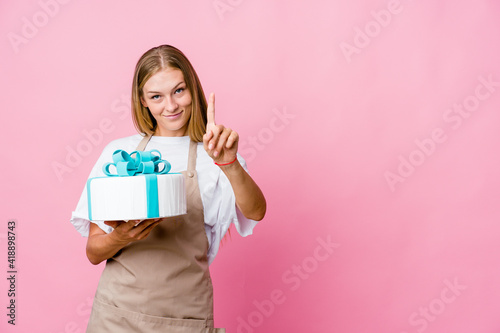 Young russian baker woman holding a delicious cake showing number one with finger.