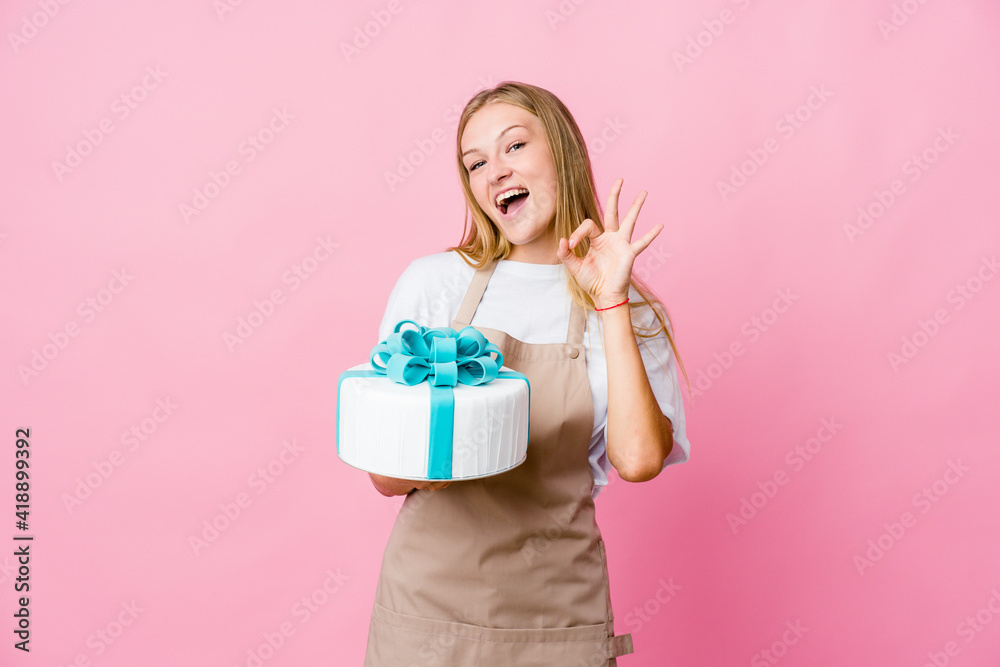 Young russian baker woman holding a delicious cake winks an eye and holds an okay gesture with hand. Young russian baker woman holding a delicious cake winks an eye and holds an okay gesture with hand.