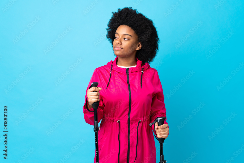 Young Africa American with backpack and trekking poles isolated on blue background making doubts gesture looking side Young Africa American with backpack and trekking poles isolated on blue background making doubts gesture looking side