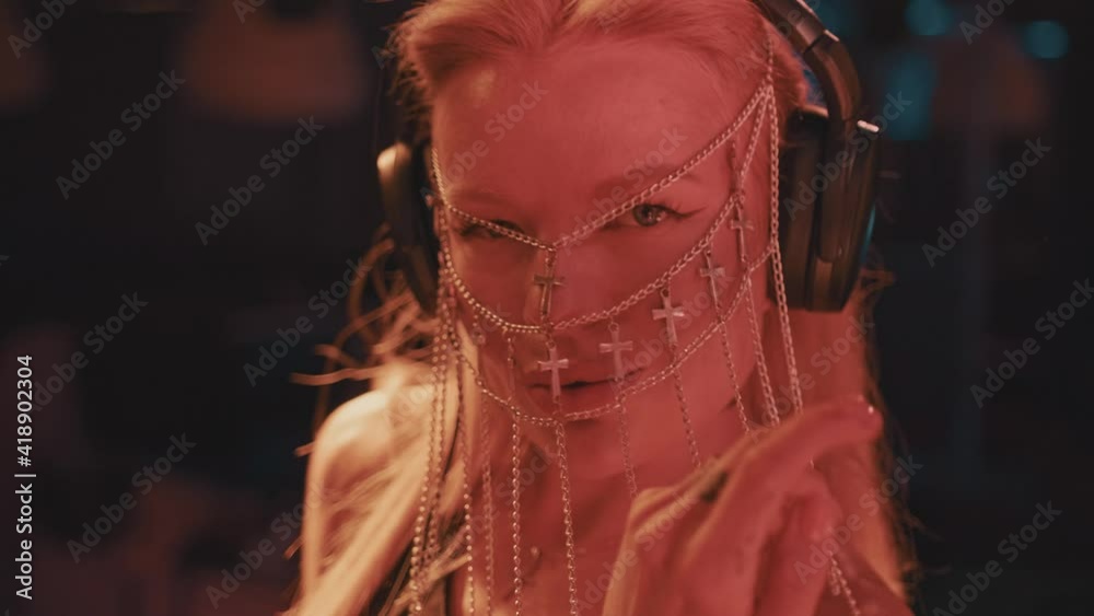 A cool and stylish female DJ of European appearance with blonde hair and a fashionable face mask made of Christian crosses works in a nightclub behind the turntables and creates dance music. A cool and stylish female DJ of European appearance with blonde hair and a fashionable face mask made of Christian crosses works in a nightclub behind the turntables and creates dance music.