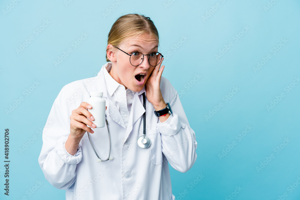 Young russian doctor woman holding pills bottle on blue shouts loud, keeps eyes opened and hands tense. Young russian doctor woman holding pills bottle on blue shouts loud, keeps eyes opened and hands tense.