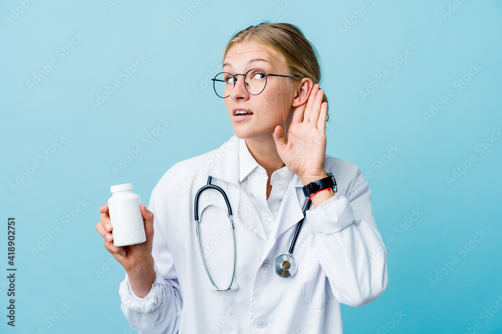 Young russian doctor woman holding pills bottle on blue trying to listening a gossip. Young russian doctor woman holding pills bottle on blue trying to listening a gossip.