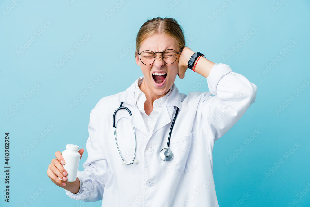 Young russian doctor woman holding pills bottle on blue covering ears with hands trying not to hear too loud sound. Young russian doctor woman holding pills bottle on blue covering ears with hands trying not to hear too loud sound.