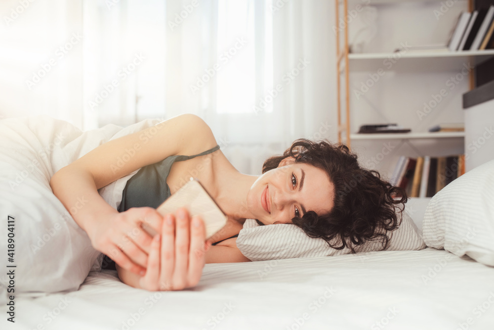 Naklejka premium Smiling woman looking at mobile phone while laying on white bed
