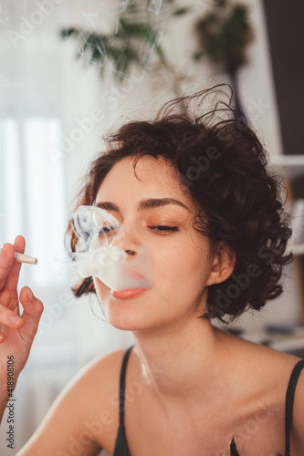 Woman sitting at her modern bedroom and smoking marijuana hand-rolled cigarettes