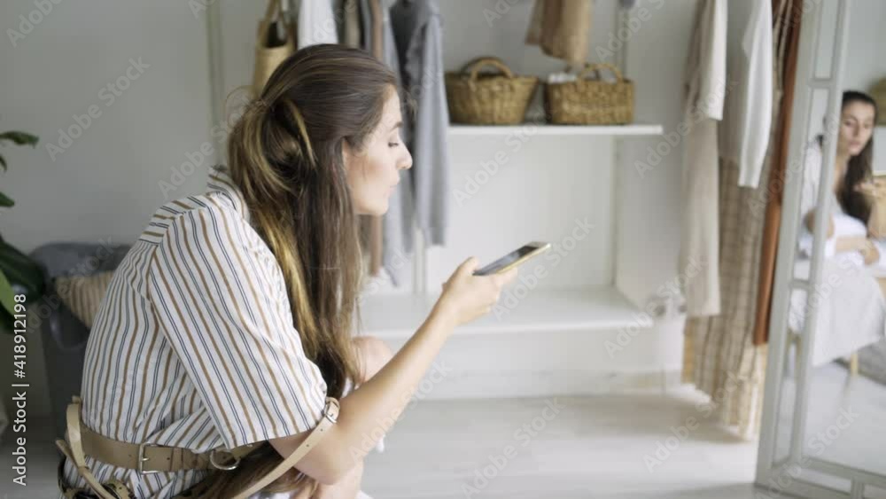 Side view elegant long haired lady in striped dress holds little baby in arms and make video call on smartphone while sit against open cupboard with hanging clothes Side view elegant long haired lady in striped dress holds little baby in arms and make video call on smartphone while sit against open cupboard with hanging clothes