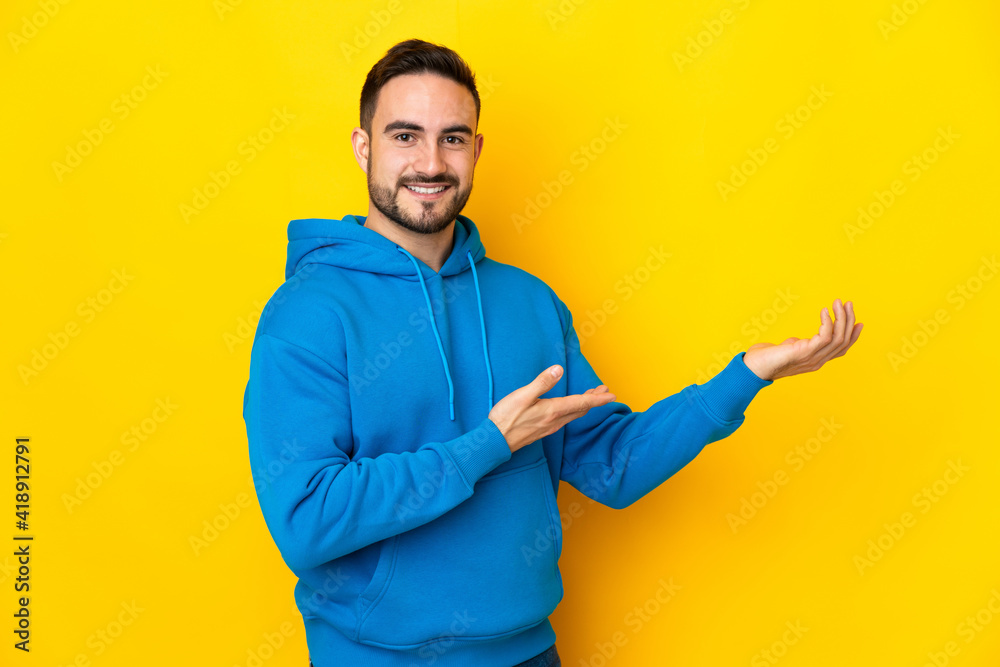 Young caucasian handsome man isolated on yellow background extending hands to the side for inviting to come Young caucasian handsome man isolated on yellow background extending hands to the side for inviting to come
