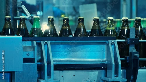 Glass bottles filled with a drink, more like beer, on the bottling line. Light illuminating bottles, movement along the conveyor line.