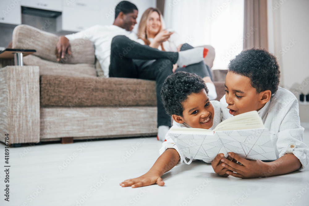 Multiracial family is together at home at daytime. Children lying down on the floor with book Multiracial family is together at home at daytime. Children lying down on the floor with book