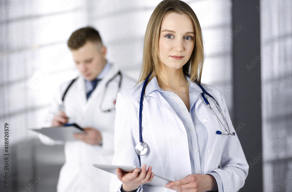 Professional woman doctor with a stethoscope is using a computer tablet, while she is standing together with her colleague in a clinic. Young doctors at work in a hospital. Medicine and healthcare Professional woman doctor with a stethoscope is using a computer tablet, while she is standing together with her colleague in a clinic. Young doctors at work in a hospital. Medicine and healthcare