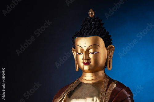 The top part of a gilded statue of Medicine Buddha, in traditional clothes with ushnisha on the head.  Isolated on black background with blue light spot.