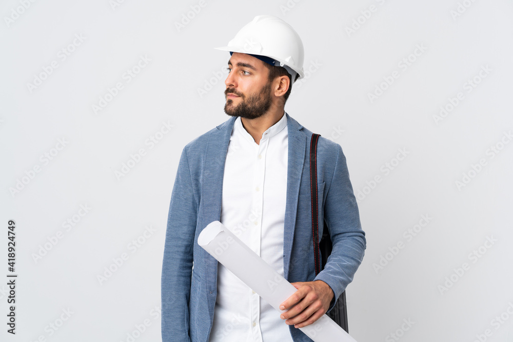 Young architect man with helmet and holding blueprints isolated on white background looking to the side - obrazy, fototapety, plakaty 