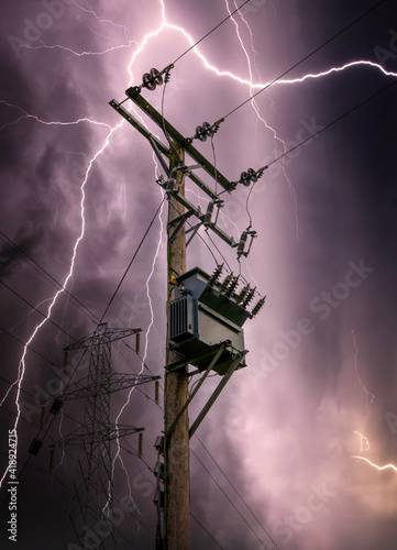 Bright lightning bolts striking electric power pylon tower cables and sub station strike. Electricity discharge cloud to ground storm with transformer on wooden telegraph pole silhouette against sky