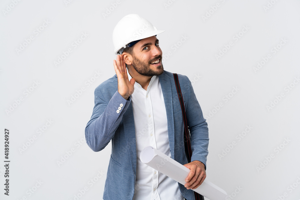 Young architect man with helmet and holding blueprints isolated on white background listening to something by putting hand on the ear