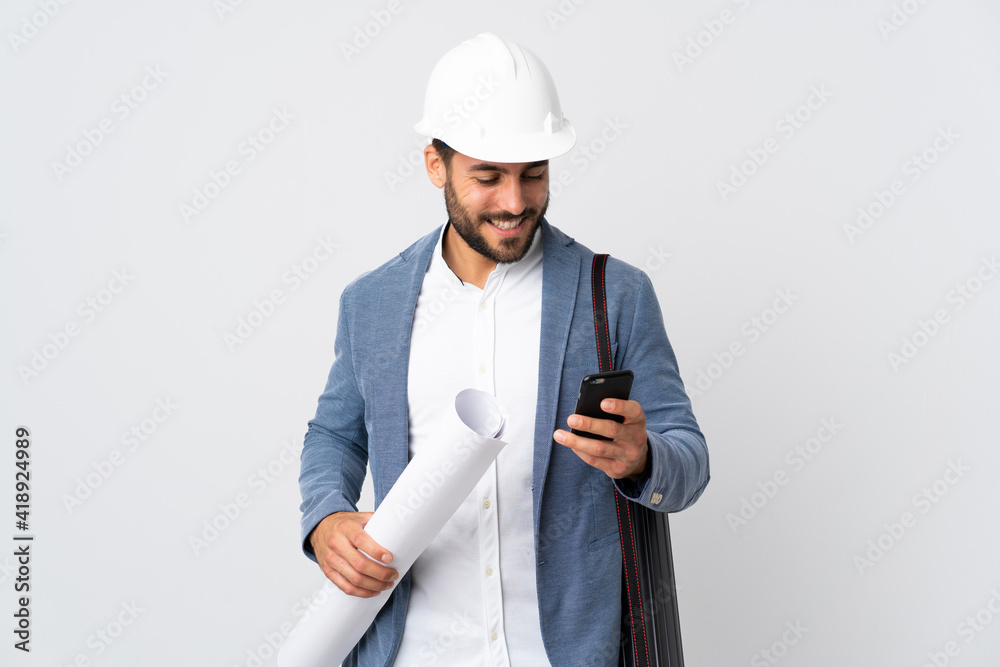 Young architect man with helmet and holding blueprints isolated on white background sending a message with the mobile Young architect man with helmet and holding blueprints isolated on white background sending a message with the mobile