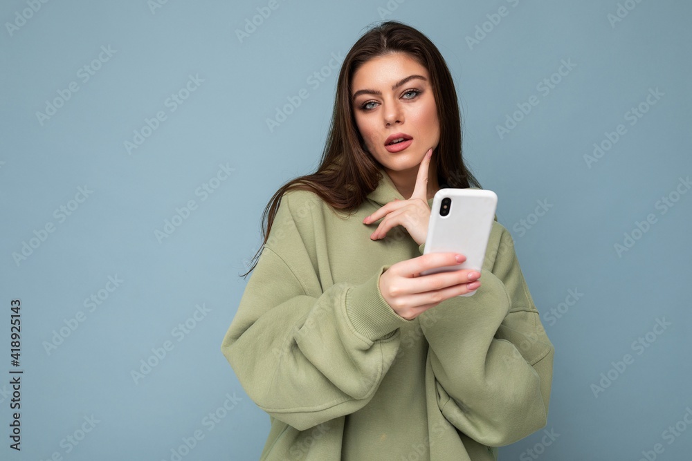 Pretty adult brunette woman wearing stylish green hoodie using mobile phone writing sms isolated on background looking at camera and thinking Pretty adult brunette woman wearing stylish green hoodie using mobile phone writing sms isolated on background looking at camera and thinking