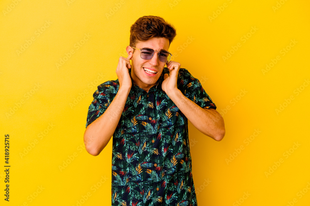 Young caucasian man wearing a Hawaiian shirt isolated on yellow background covering ears with hands.