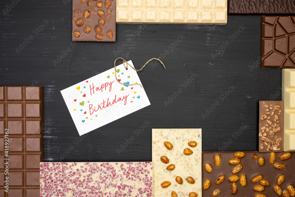 Obraz premium different sweet, delicate chocolate bars are creative on a black wooden background with a label that says happy birthday