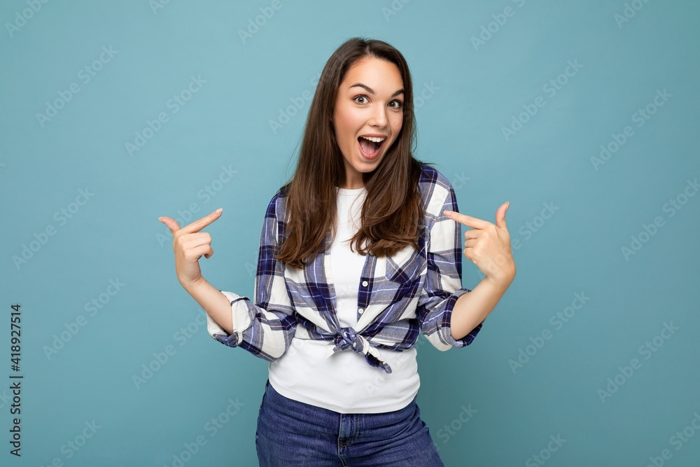 Portrait of young positive happy attractive amazing beautiful brunette woman with sincere emotions wearing hipster blue check shirt isolated over blue background with copy space and pointing at Portrait of young positive happy attractive amazing beautiful brunette woman with sincere emotions wearing hipster blue check shirt isolated over blue background with copy space and pointing at