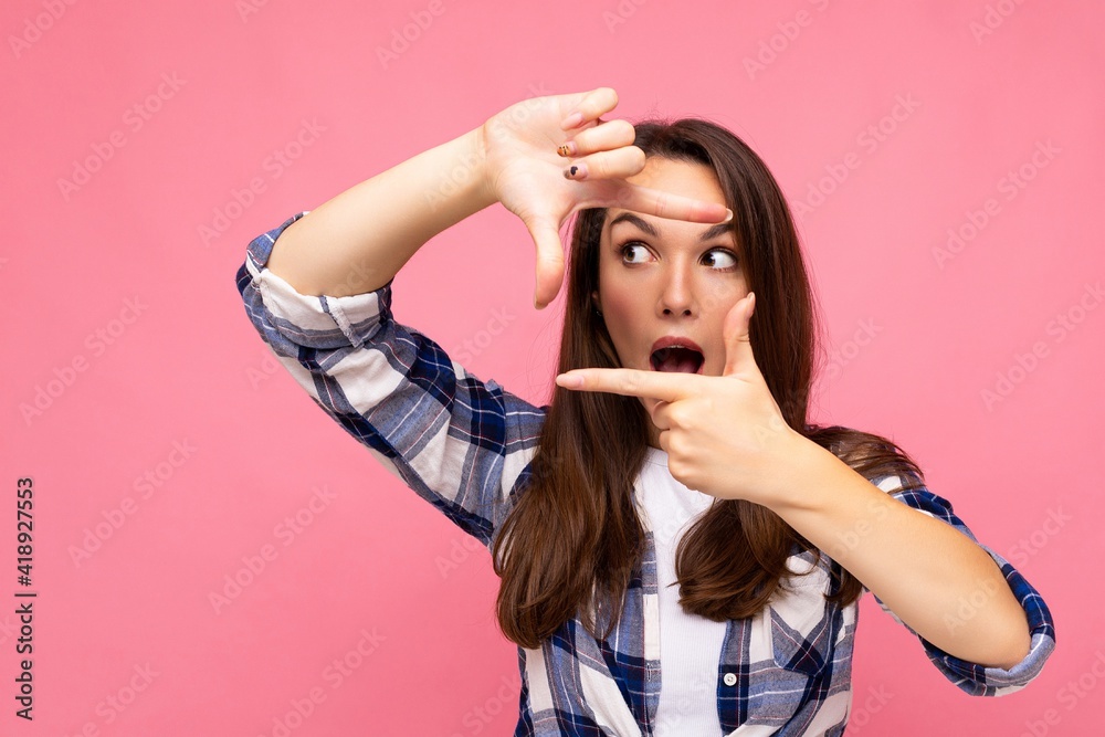 Fototapeta premium Young positive delightful amazed pretty brunette woman with sincere emotions wearing hipster check shirt making frame with hands, taking picture with imaginary camera and standing isolated on pink