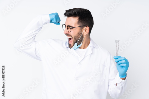 Dentist man holding tools isolated on white background celebrating a victory