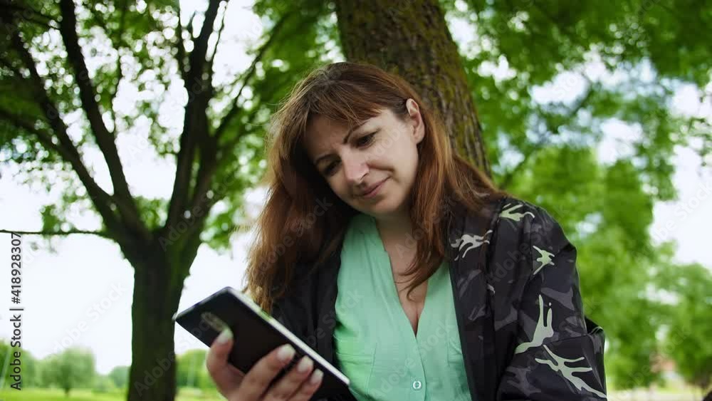 brunette woman sits in the park under a tree uses the phone and smiling, read some information, chat with friends brunette woman sits in the park under a tree uses the phone and smiling, read some information, chat with friends