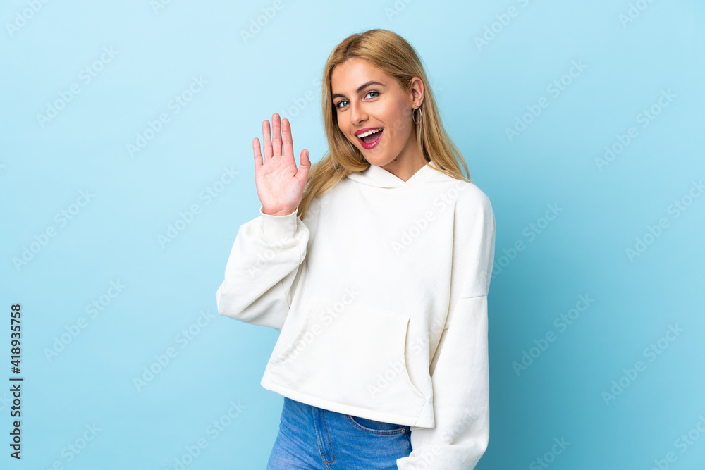 Fototapeta premium Young Uruguayan blonde woman over isolated blue background saluting with hand with happy expression