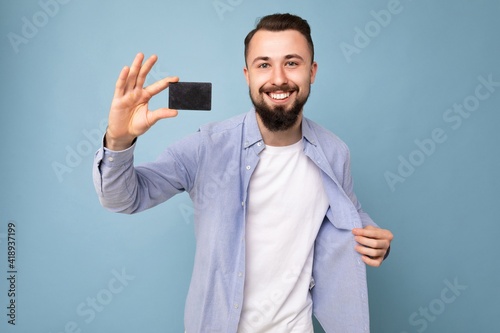 photo shot of Handsome smiling brunette bearded young man wearing stylish blue shirt and white t-shirt isolated over blue background wall holding credit card looking at camera