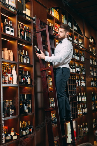 The seller of alcoholic beverages sorts bottles standing on the ladder. © davit85