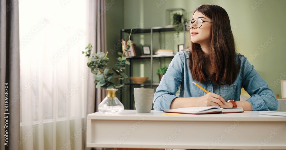 Young beautiful student girl with glasses sitting at desk in apartment and writes into the diary task for week. Young beautiful student girl with glasses sitting at desk in apartment and writes into the diary task for week.