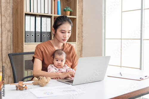 Busy woman trying to work while babysitting newborn baby daughte.  Young beautiful Asian mother holding little baby on lap while another hand typing. Working women with multitasking.