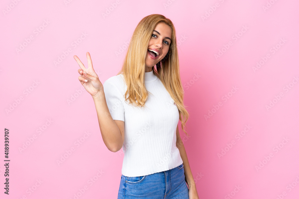 Young Uruguayan blonde woman over isolated pink background smiling and showing victory sign Young Uruguayan blonde woman over isolated pink background smiling and showing victory sign