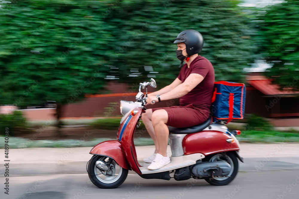 Delivery boy wearing red uniform on scooter with isothermal food case box driving fast. Express food delivery service from cafes and restaurants. Courier on the moto scooter delivering food.