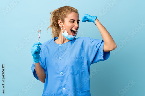 Young woman dentist holding tools over isolated blue background celebrating a victory