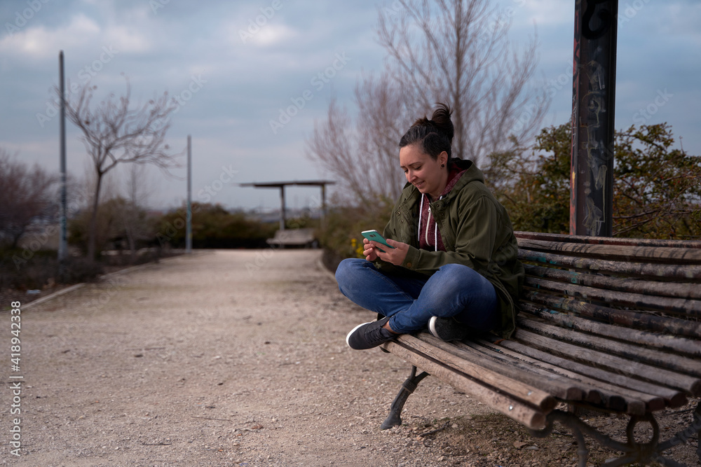 Fototapeta premium Young woman sitting on a park bench texting with her phone