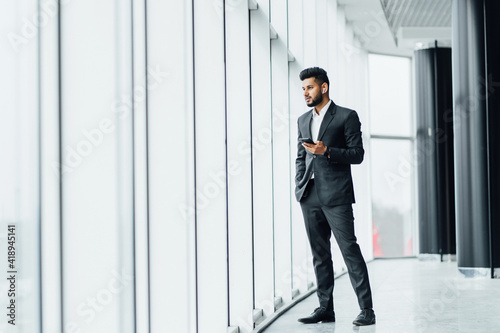 A young Indian businessman in a modern office building, he is standing by the window and talking on the phone, a business meeting, wearing a black suit