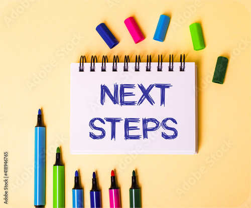 The words NEXT STEPS written in a white notebook on a beige background near multi-colored markers photo