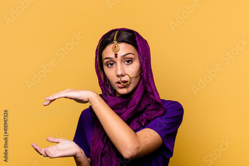 Young Indian woman wearing a traditional sari clothes isolated on yellow background shocked and amazed holding a copy space between hands. © Asier