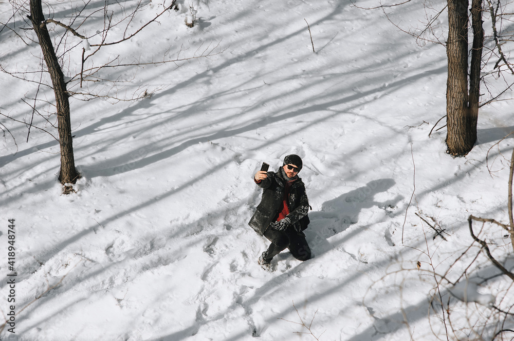 A man, a wanderer, a tourist with a large black backpack on his back travels through a snowy forest in countries and takes pictures of the beautiful nature and himself on a mobile phone, smartphone.