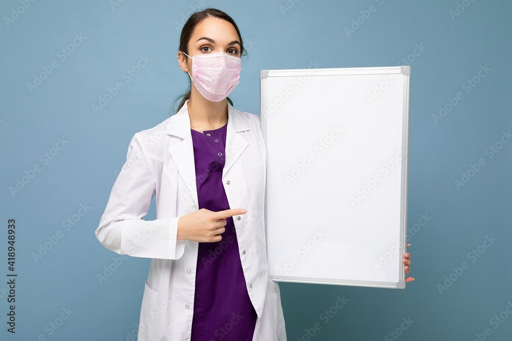 Nurse in protective face mask and white medical coat holding an empty magnetic board isolated on blue background Nurse in protective face mask and white medical coat holding an empty magnetic board isolated on blue background