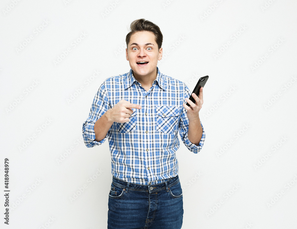 Young excited man in blue shirt pointing finger to blank screen of phone to viewer with WOW face